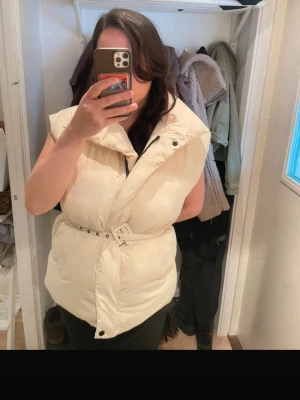 Krämvit dunväst med bälte - Snygg krämvit dunväst med bred krage och tillhörande bälte i midjan. Västen har en quiltad design och stängs med både dragkedja och tryckknappar. Perfekt för lager-på-lager och ger en trendig siluett.