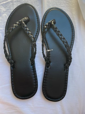 Svarta flätade sandaler - Snygga svarta sandaler med flätade remmar och klassisk flip-flop design. Råkade beställa två drycker så dessa är aldrig använda 