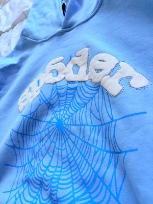 Blå Sp5der hoodie med spindelnät - Säljer en ljusblå hoodie från Sp5der med ett stort blått spindelnätsmotiv på bröstet och vit 3D-text. Hoodien har huva och är tillverkad i mjuk bomullsblandning. Perfekt för dig som gillar streetwear och vill sticka ut med en unik design.