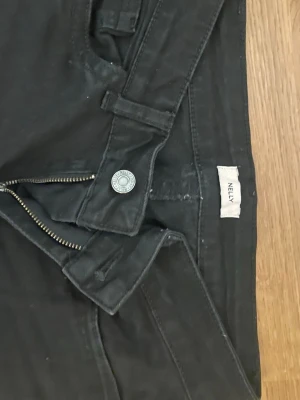 Svarta jeans från Nelly - Säljer ett par svarta jeans från Nelly med detaljer på bakfickorna. Jag köpte dom men de har bara använts 4 gånger innan dom glömdes bort i garderoben och inte kommit till användning. De är inte slitna och har inga skador. Storlek 36.