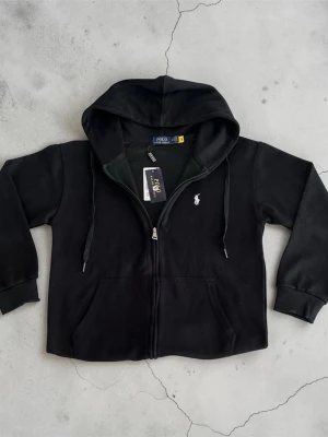 Svart hoodie från Polo Ralph Lauren storlek M (kännst som S) - Svart hoodie med dragkedja från Polo Ralph Lauren. Klassisk design med huva, dragsnören och två fickor framtill. Liten vit broderad logga på bröstet. Tillverkad i mjukt bomullsmaterial för en skön känsla. M (men kännst som S)