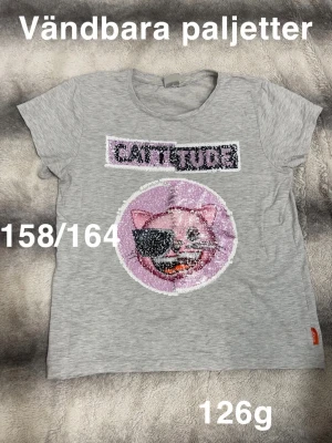 Grå t-shirt med vändbara paljetter - Grå t-shirt med coolt kattmotiv och texten 'CATTITUDE' i vändbara paljetter. Rund halsringning och korta ärmar. Perfekt för dig som gillar unika detaljer och glitter.🌸 Fint använt skick. Vändbara paljetter. Storlek 158/164 