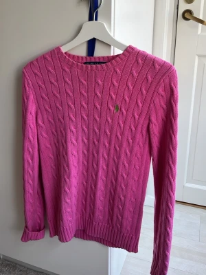 Rosa kabelstickad tröja Polo Ralph Lauren - Rosa kabelstickad tröja från Polo Ralph Lauren med rund halsringning och grönt broderat logotyp på bröstet. Tröjan har långa ärmar och ribbstickade muddar vid ärmslut och nederkant. Perfekt för dig som gillar klassisk och färgglad stil. M men jag som brukar ha xxs passar den ändå bara lite lång i armarna! Använd få gånger!