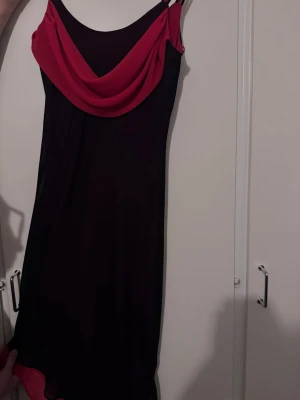 Svart och röd klänning med drapering - Svart klänning med röda detaljer och draperad urringning. Klänningen har smala axelband och en röd volangkant nertill. Snygg och figurnära modell som ger en elegant look.