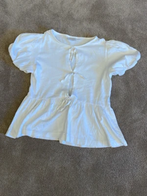 Vit blus med puffärm och knyt - Söt vit blus med puffärmar och tre dekorativa knyt framtill. Blusen har en lätt volang nedtill och är tillverkad i ett mjukt bomullstyg. Perfekt för en trendig och look! Har använt dessa ett förtal gånger bara! 