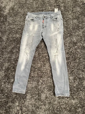 Ljusgrå slitna jeans från Dsquared2 - Säljer ett par ljusgråa jeans från Dsquared2 med skinny fit och slitna detaljer på benen med gröna färgfläckar. Jeansen har klassisk femficksmodell, knappgylf och tydliga slitningar för en trendig look. Perfekta för dig som gillar streetstyle och vill sticka ut. Byxorna är M/Lmen är ganska stora i midjan rekommenderar bälte, byxorna har en ganska bra sick 