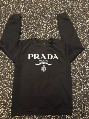 Svart sweatshirt från Prada - Svart sweatshirt med rund hals och tryck framtill med texten 'PRADA MILANO DAL 1913' samt emblem. Tröjan har långa ärmar och ribbade muddar vid ärmslut och nederkant. Perfekt för en stilren och trendig look. Passar också S eftersom den är liten i storlek