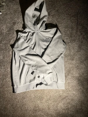 Grå Peak Performance hoodie med dragkedja - Snygg grå hoodie från Peak Performance med dragkedja framtill och broderad logga på bröstet. Mjuk insida och klassisk huva. Perfekt för chill dagar eller när du vill ha en clean och enkel stil.