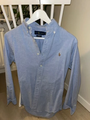 Blå skjorta från Ralph Lauren - Klassisk ljusblå skjorta från Ralph Lauren med button-down krage och broderad logga på bröstet. Skjortan har långa ärmar och vita knappar framtill. Tillverkad i mjuk bomull för en bekväm känsla.