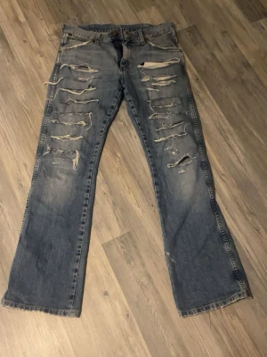 Blå slitna jeans med raka ben - Säljer ett par blå jeans med raka ben och kraftigt slitna detaljer framtill och baktill. Jeansen har klassisk femficksdesign och tydliga slitningar och hål för en edgy look. Perfekt för dig som gillar streetstyle och avslappnad vibe.