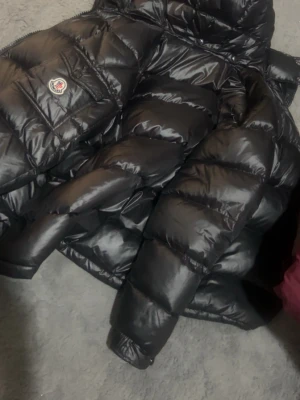 Svart Moncler dunjacka med huva - Säljer en svart Moncler dunjacka med huva och stora quiltade partier. Jackan har dragkedja, ficka med Moncler-logga på ärmen och coolt serietryck på insidan. Perfekt för kalla dagar och riktigt snygg streetstil.. den går att skanna. Storlek S, pris går diskuteras