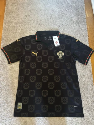 Svart Portugal fotbollströja Puma - Snygg svart Portugal fotbollströja från Puma med gulddetaljer och mönster av Portugals landslagssköld över hela tröjan. Krage med rödgröna detaljer och Puma-logga på bröstet. Tillverkad i lätt, andningsbart material med dryCELL-teknologi.