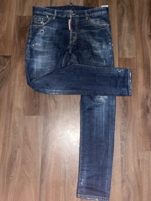 Ds2 Jeans - Snygga mörkblå slim fit jeans från Dsquared2 med slitningar, färgstänk och klassisk femficksdesign. Jeansen har en låg midja och smal passform, samt detaljerade sömmar och en röd logotagg vid gylfen. Perfekta för en trendig streetstil.