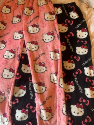 Hello Kitty loungewear byxor 2-pack - Två par mjuka loungewearbyxor i fleece med Hello Kitty-motiv. Ena paret är rosa med vita Hello Kitty-ansikten och röda rosetter, det andra är svart med samma mönster. Perfekta för mysiga hemmakvällar och chill.