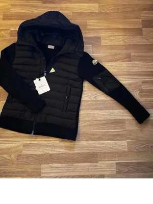 Svart dunväst med huva från Moncler - Snygg svart dunväst från Moncler med huva och dragkedja framtill. Västen har stickade ärmar, två sidofickor med dragkedja och Moncler-logga på ena ärmen. Perfekt för lager-på-lager och streetstyle. Material: dunfyllning och stickade detaljer.