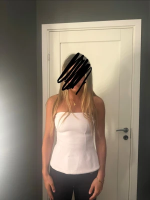 Vit bandeau tubtopp - Säljer en stilren vit bandeau tubtopp med figurnära passform. Toppen är axelbandslös och har en enkel design med sömmar som markerar formen. Perfekt att matcha med jeans eller kjol för en clean look.