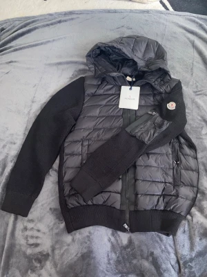 Svart cardigan från Moncler - Helt ny Cardigan från Moncler med stickade ärmar och vadderad kropp. Jackan har huva, dragkedja framtill och två fickor. Moncler-logga på ena ärmen. Perfekt för kalla vinterdagar och riktigt trendig look. Priset kan diskuteras.