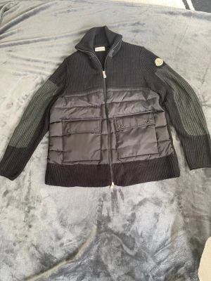 Svart cardiganjacka från Moncler - Helt ny svart cardiganjacka från Moncler med stickade ärmar och krage samt vadderad front. Jackan har två stora fickor med lock och dragkedja framtill. Klassisk Moncler-logga på ärmen. Priset kan diskuteras.