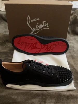 Christian Louboutin svarta sneakers - Svarta sneakers från Christian Louboutin med ikoniska röda sulor och svarta nitar på tån. Skorna har lågt skaft, snörning och är tillverkade i läder och textil. Perfekt för dig som vill sticka ut med en lyxig och edgy stil.