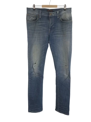 Nudie Jeans Co Thin Finn Blue Stone Jeans, W33 L34 - Ett par Nudie Jeans Co. jeans i en ljusblå tvätt. Jeansen har en dragkedja framtill och fickor både fram och bak. Designad med trendiga hål på knäna. Midja: 44cm Långt: 81cm Benöppning: 18cm