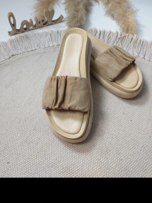 Beige sandaler från Indora x Nicole - Säljer ett par trendiga beige sandaler från Indora x Nicole i läder, med breda, rynkade remmar i mocka och tjocka sulor. Perfekta för dig som gillar en avslappnad och stilren look. Materialet är mjukt och bekvämt, och designen är enkel men modern.
