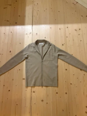 Beige ziptröja från Samsøe Samsøe - Stilren beige ziptröja från Samsøe Samsøe med lång ärm, krage och bröstficka. Tröjan har en rak passform och är tillverkad i ett mjukt material som känns skönt mot huden. Perfekt för lager på lager och enkel att matcha.