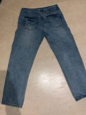 Blå mom jeans i klassisk modell - Säljer ett par blå mom jeans med hög midja och raka ben. Jeansen har fem fickor, bälteshällor och stängs med dragkedja och knapp. Materialet är klassisk denim i bomull och färgen är ljusblå med lätt tvättade detaljer.