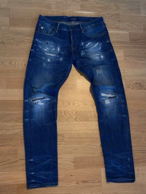 Blåa jeans distressed med randig bakficka - Säljer ett par blå jeans från Scotch & Soda med slitningar och trasiga detaljer på framsidan. Bakfickan har ett unikt randigt tyg som sticker ut. Jeansen har smal passform och klassisk femficksdesign. Perfekt för dig som gillar en edgy och trendig look.