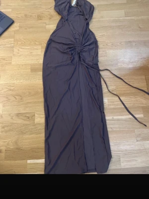 Brun maxiklänning med pärldetaljer - Säljer en brun maxiklänning med asymmetrisk axel och dekorativa pärlor framtill. Klänningen har dragsko i midjan och långa band som kan knytas. Materialet är mjukt och följsamt, perfekt för en trendig och unik look.