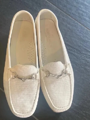 Vita loafers med metallspänne - Säljer ett par stilrena vita loafers med dekorativt metallspänne framtill. Skorna har en klassisk fyrkantig tå och är tillverkade i ett texturerat skinnmaterial. Perfekta för dig som gillar en clean och tidlös look.