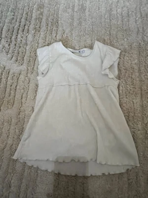 Vit kortärmad topp med volangärm - Söt vit topp med korta volangärmar och rund halsringning. Toppen har en lätt utsvängd passform och vågig kant nertill. Perfekt för en avslappnad och trendig look.