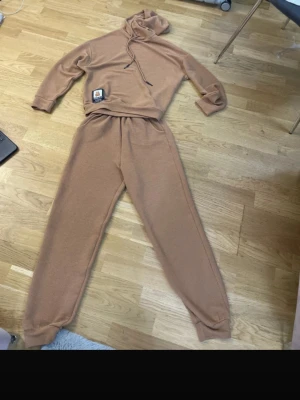 Brun ribbad loungewear-set med luva - Mysigt loungewear-set i brun ribbad trikå med hoodie och matchande byxor. Hoodien har dragsko och en söt nalle-patch framtill. Byxorna har resår i midjan och muddar vid benslut. Perfekt för chill dagar hemma.