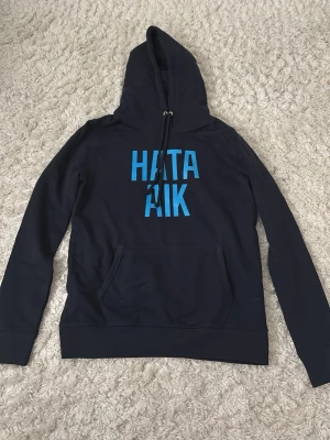 Svart hoodie med blå text HATA AIK - Svart hoodie med huva och känguruficka framtill. Tryck i blått med texten 'HATA AIK' på bröstet. Klassisk modell med ribbade muddar vid ärmslut och nederkant. Perfekt för dig som vill sticka ut med ett tydligt statement.