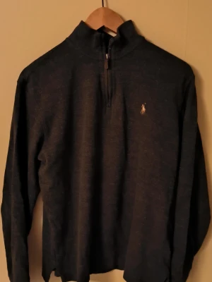 Svart stickad tröja Polo Ralph Lauren - Svart stickad tröja från Polo Ralph Lauren med halv dragkedja och klassisk broderad logga på bröstet. Tröjan har lång ärm och hög krage, perfekt för lager på lager. Materialet känns mjukt och bekvämt. Priset är ej hugget i sten