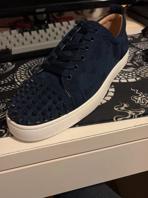 Blåa skor från Christian Louboutin med spikar - Snygga blåa sneakers från Christian Louboutin med mockaöverdrag och coola svarta nitar på tån. Skorna har klassiska vita sulor och den ikoniska röda undersidan med Louboutin-loggan. Perfekt för dig som vill sticka ut med exklusiva detaljer.