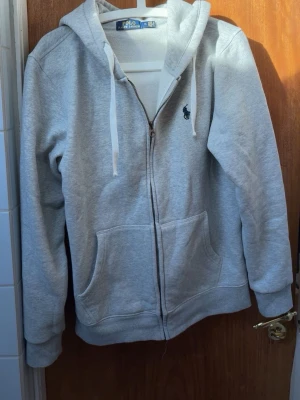 Ralph Lauren zip hoodie - Snygg grå zip hoodie från Polo Ralph Lauren med klassisk marinblå logga broderad på bröstet. Hoodien har vita dragsnören, två stora fickor framtill och ribbade muddar. 