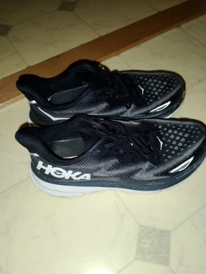 Svarta Hoka Clifton 9 strl 42.5 - Säljer ett par svarta Hoka sneakers med vit logga på sidan och detaljer i vitt. Ovandelen är i ventilerande mesh och sulan är tjock och dämpande för extra komfort. Klassisk låg modell med snörning, perfekt för dig som gillar sportig stil. Väldigt bekväma att använda för fötterna. Storlek 42.5. använda sparsamt , i mkt bra skick ! Nypris runt 2000:-