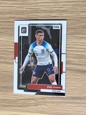 FIFA samlarkort Phil Foden - Samlarkort från Panini Donruss Soccer Optic 2022-23 med Phil Foden i Englands landslagströja. Kortet har färgerna vitt, blått och rött och visar spelaren i matchställ. Perfekt för dig som gillar fotboll och samlar på sportkort.