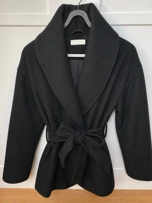 Svart kappa - Stilren svart kappa med bred sjalkrage och knytskärp i midjan. Kappan har en avslappnad passform och är tillverkad i ett mjukt tyg som ger en lyxig känsla. Kappan är från Gina tricot premium. 