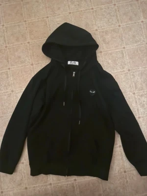 Svart Comme des Garçons PLAY hoodie - Svart hoodie med dragkedja från Comme des Garçons PLAY. Ikoniskt hjärta med ögon på bröstet och stort hjärta med ögon på ryggen. Tillverkad i mjukt bomullsmaterial och har justerbar huva samt fickor framtill. Perfekt för en streetwear-look.