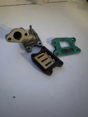 Insugsdelar till moped - Säljer ett kit med insugsdelar till moped, inklusive insug, reedventil och packning. Delarna är i metall och plast, med insuget i silverfärgad metall och reedventilen i svart plast med metallblad. Perfekt för dig som vill trimma eller byta ut delar på din moppe.