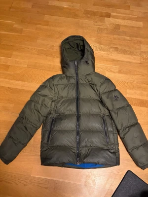 Olivgrön dunjacka från J.Lindeberg - Snygg olivgrön dunjacka från J.Lindeberg med huva och två stora dragkedjeförsedda fickor framtill. Jackan har en quiltad design och ett tydligt J.Lindeberg-märke på ärmen. Perfekt för kalla vinterdagar och riktigt varm.