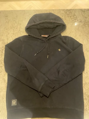 Svart hoodie från Morris Stockholm - Svart hoodie från Morris Stockholm med dragsko i huvan och liten guldfärgad logga på bröstet. Mjuk bomullskänsla och ribbade muddar vid ärmslut och nederkant. Märkespatch nertill på framsidan. Perfekt för en chill och stilren look.