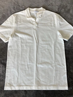 Vit t-shirt med krage från Bläck - Stilren vit t-shirt från Bläck med korta ärmar och unik krage utan knappar. Tillverkad i mjuk bomull som känns skön mot huden. Perfekt för en clean och enkel look.