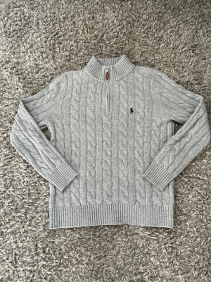 Beige kabelstickad tröja Polo Ralph Lauren - Kabelstickad beige tröja från Polo Ralph Lauren med halv dragkedja och broderad logga på bröstet. Tröjan har ribbade muddar och krage, samt klassisk kabelstickad design. Perfekt för lager-på-lager och snygg till jeans.