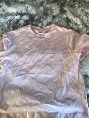 Ljusrosa t-shirt från Gina Tricot - En basic ljusrosa t-shirt från Gina Tricot, basically basics-kollektionen. T-shirten har rund halsringning och korta ärmar. Perfekt för en enkel och stilren look. Tillverkad i mjuk bomull för maximal komfort.