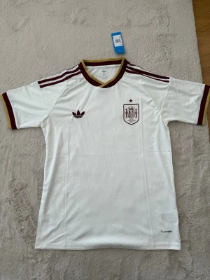 Vit Adidas Spanien fotbollströja - Säljer en vit fotbollströja från Adidas med Spaniens landslagsmärke på bröstet. Tröjan har vinröda detaljer på krage, ärmslut och axlar samt Adidas-logga. Materialet är lätt och andas, perfekt för träning eller match.
