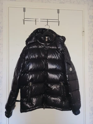 Svart pufferjacka från Moncler - Säljer en svart, glansig pufferjacka från Moncler med huva och dragkedja framtill. Jackan har två framfickor med dragkedja och Moncler-logga på ärmen. Perfekt för kalla vinterdagar och riktigt snygg streetstil.