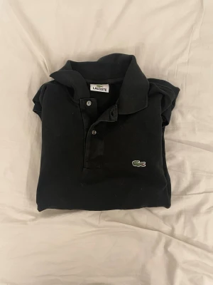 Svart pikétopp från Lacoste - Svart pikétopp från Lacoste med klassisk krage, knappar framtill och den ikoniska gröna krokodilloggan på bröstet. Tillverkad i mjuk bomull för en bekväm känsla. Enkel och stilren design som funkar till det mesta.