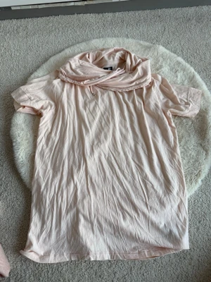 Ljusrosa topp med stor krage från JC - Ljusrosa topp från JC med korta ärmar och en stor, mysig krage som kan stylas på olika sätt. T-shirten är gjord i mjuk bomull och har en relaxed passform. Perfekt för dig som gillar en enkel men unik look.